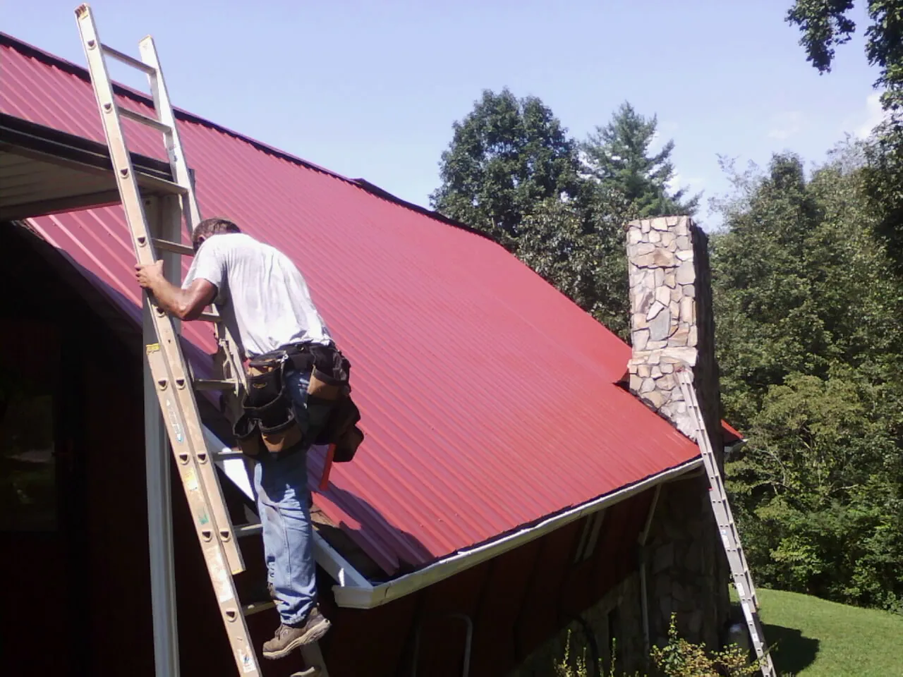 Roofer on a ladder beside a red metal roof and stone chimney — Soffit & Fascia Repair by Roofing Brothers in Ottumwa